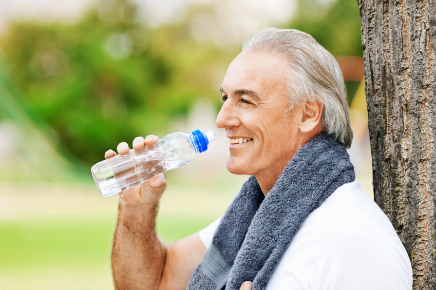 Senior man takes a drink from his water bottle after exercising in the park. He has a towel draped around his neck. Horizontal shot.