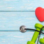 A heart with a stethoscope lies on a wooden background next to dumbbells. Healthy heart; Heart-Healthy Exercises