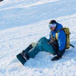 A smiling male snowboarder resting sit against a blue sky.