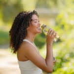 Brazilian fitness lady enjoying water drinking from bottle outdoors, posing happily in sportswear, ready for morning training in nature. Health and sport motivation.