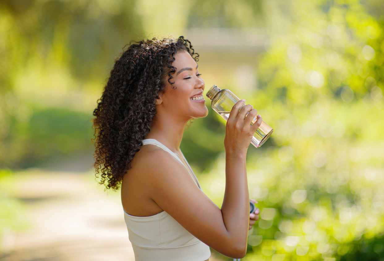 Brazilian fitness lady enjoying water drinking from bottle outdoors, posing happily in sportswear, ready for morning training in nature. Health and sport motivation.
