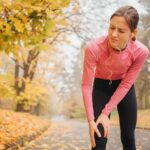 Slim and well-built young woman stands on road in autumn park. She holds hands on knee in pain.