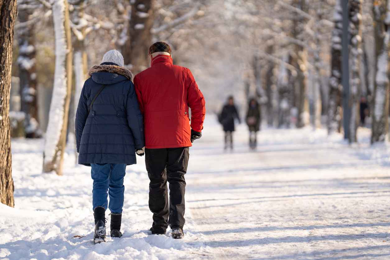 rear view of senior couple dressed in warm clothes walking on snow covered avenue in park of a city on cold sunny winter day